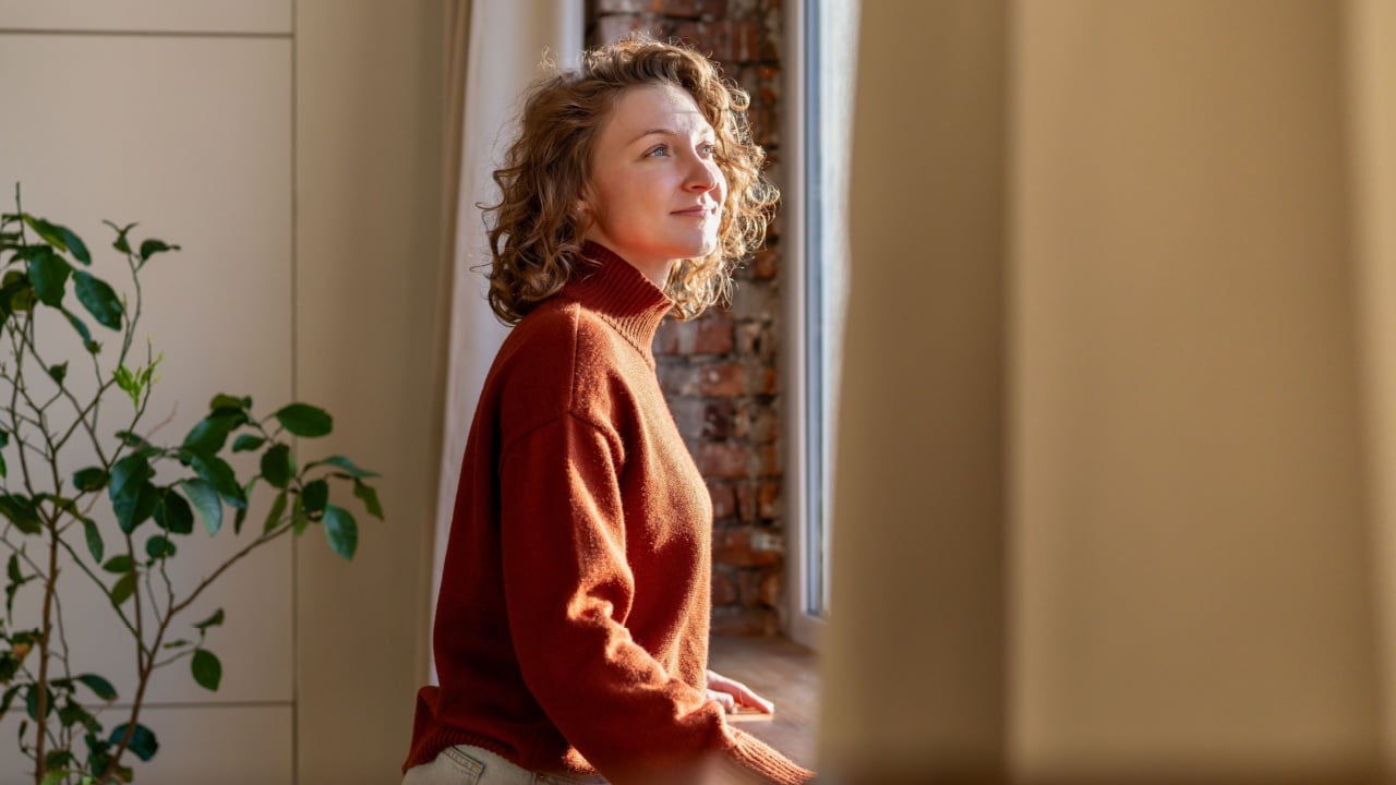 Positive thoughtful woman enjoys tranquil break at home, leaning by window feeling gentle sunlight on skin. Carefree young female with peaceful heart, joyful mind relishes beauty of day taking pause.