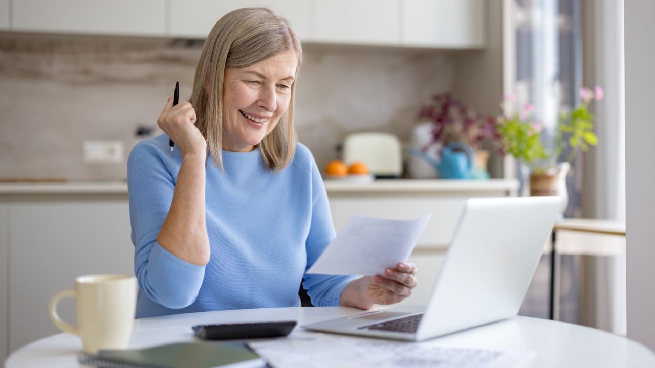 Senior woman smiling at kitchen table, reviewing bills and documents while using a laptop to budget, manage savings and plan a secure, confident retirement future